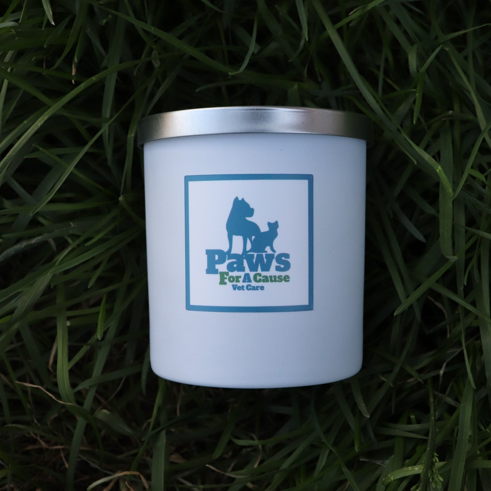 AURA Candle Bar's PAWS For A Cause Vet Care candle in a matte white vessel with a silver lid.  Candle vessel is laying on a bed of soft green grass.  